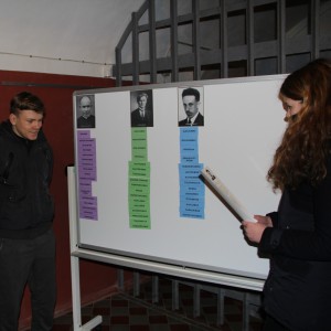 Du moksleiviai stovi prie lentos. Mergina rankoje laiko popieriaus lapus su tekstu. Ant lentos – trijų žydų gelbėtojų fotografijos, o po jomis priklijuoti lapeliai su asmenybes apibūdinančiais žodžiais.