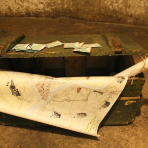 Pravira žalios spalvos medinė dėžė, ant kurios padėta daug nedidelių IX forto gyvybinės sienos žemėlapių. Iš dėžės kyšo Kauno žemėlapis.